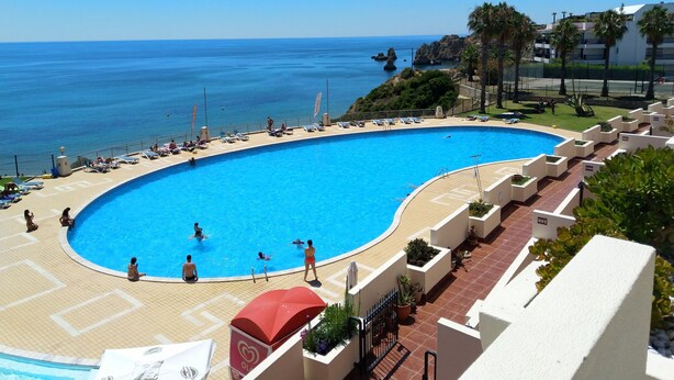A large outdoor swimming pool with a curved edge, surrounded by lounge chairs and umbrellas, overlooking a clear blue sea. - Casa Pimenta near the beach, Iberlagos Resort (Lagos)