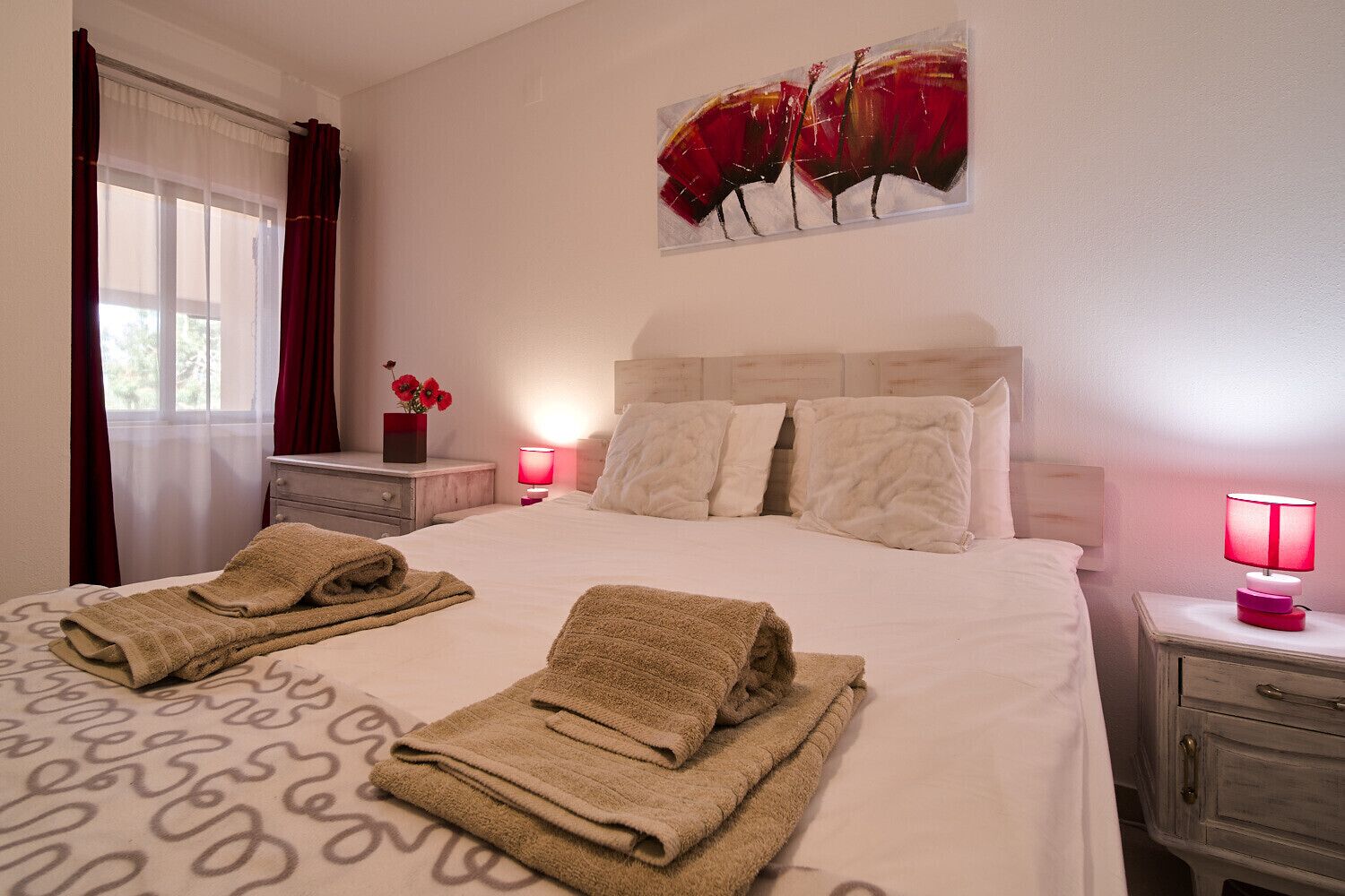 Una cama bien hecha con dos toallas dobladas, una mesita de noche con una lámpara y un cuadro en la pared.