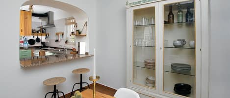 A dining area with a wooden table, three stools, a glass cabinet with dishes, and a kitchen visible through an archway.