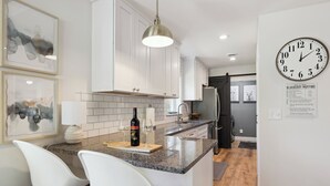 Una cocina moderna con armarios blancos, encimera de granito y refrigerador de acero inoxidable. Hay dos taburetes blancos de bar y una lámpara colgante.