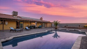 A swimming pool with lounge chairs and a patio area, surrounded by a building with a tiled roof and a clear sky.