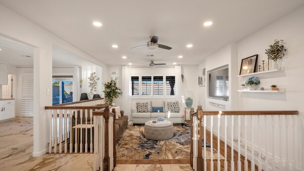 A spacious living area with a marble floor, a central seating area with a sofa and ottoman, a ceiling fan, and a large window with blinds.