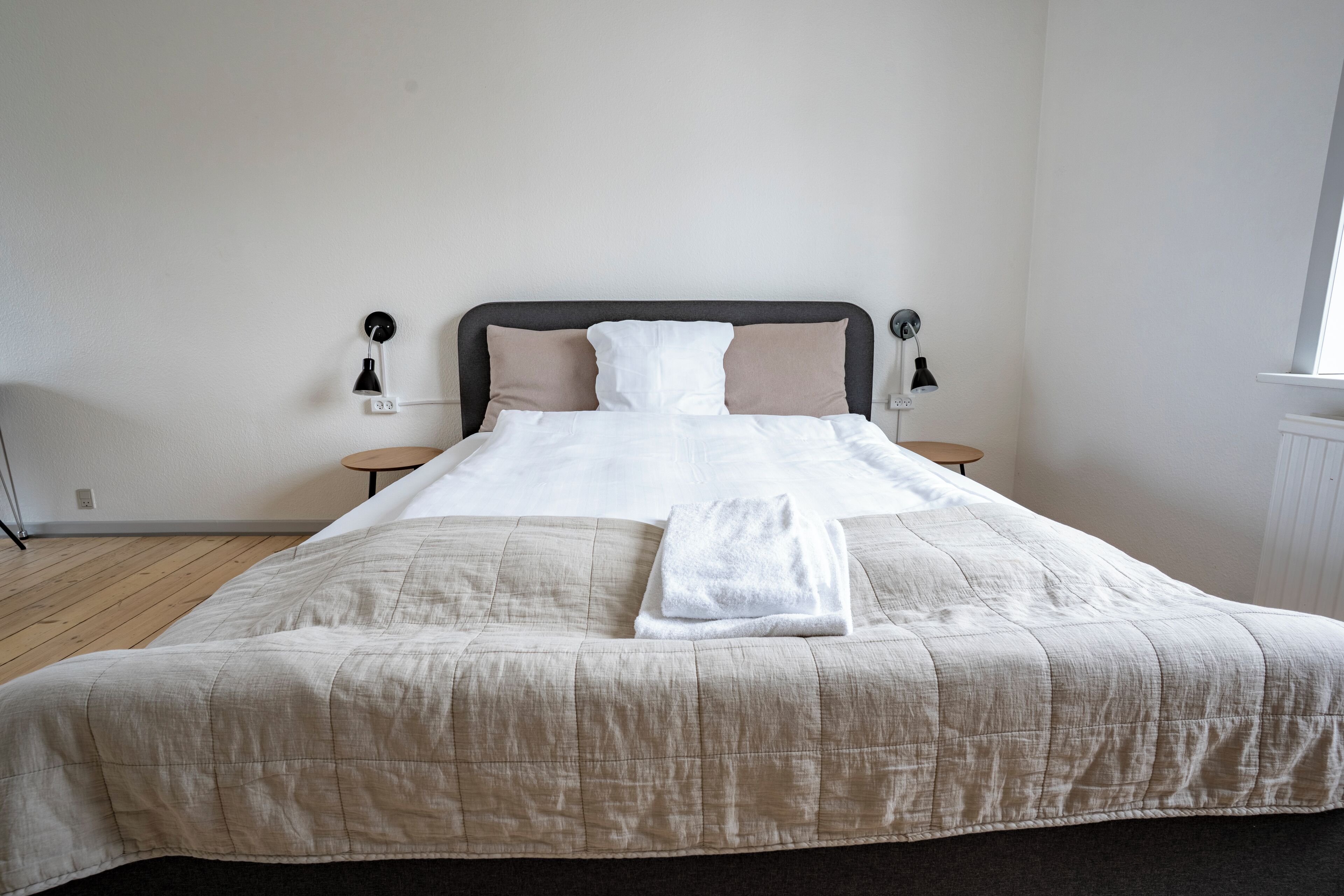 A neatly made bed with a beige quilt, two bedside tables with lamps, and a window on the right.