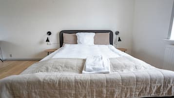 Una cama bien hecha con un edredón color beige, dos mesitas de noche con lámparas, y una ventana a la derecha.