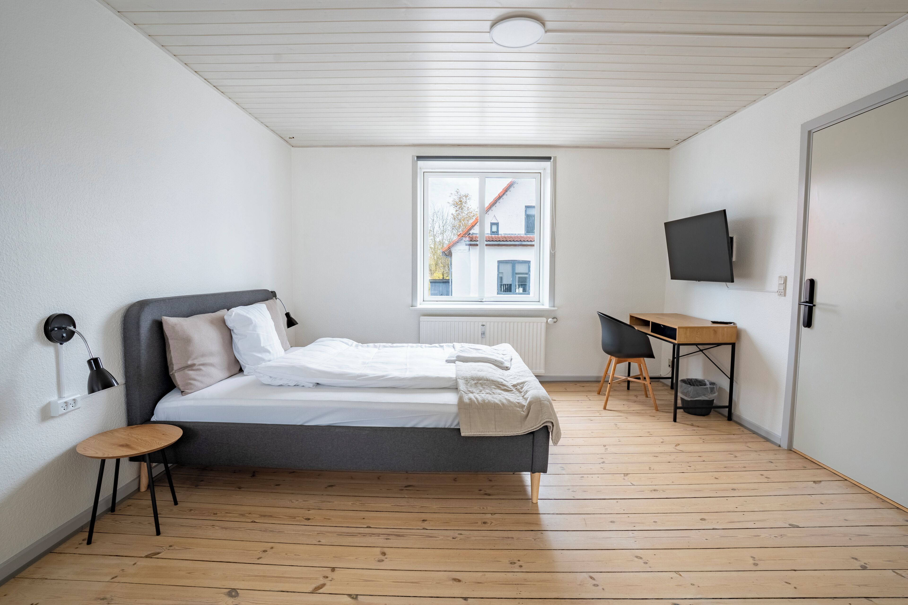 Une chambre à coucher comprenant un lit, un bureau, une chaise, un téléviseur et une fenêtre donnant sur des immeubles.