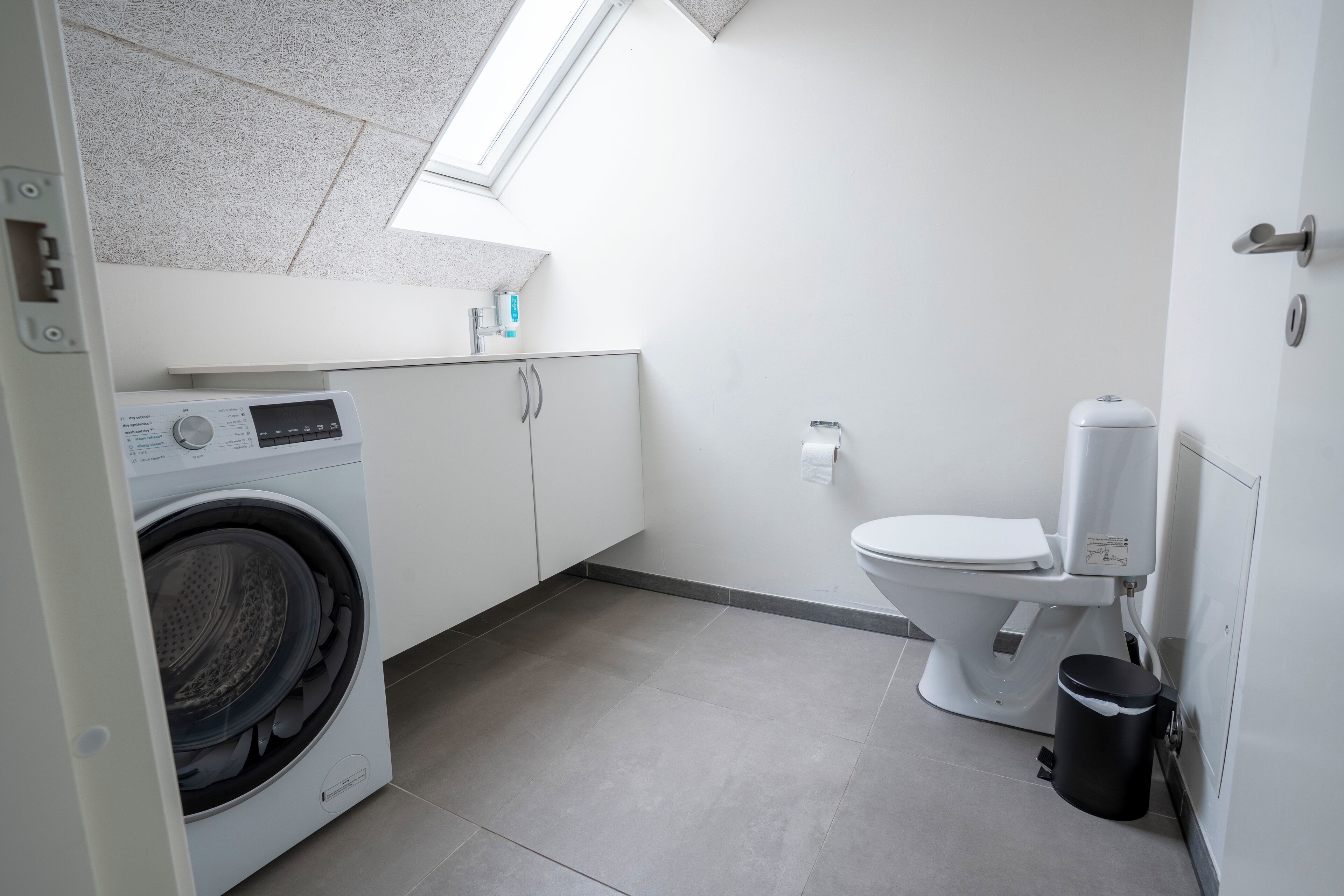 A compact, modern bathroom with a built-in washing machine, a toilet, and a trash bin.