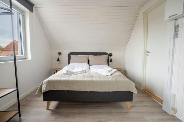 A bedroom with a sloped ceiling, a bed with a beige quilt, two bedside tables, and a window with a view of buildings. - City Apartments (Holstebro)
