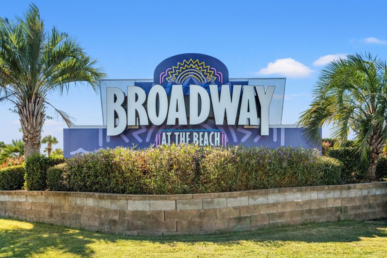 A sign for 'Broadway at the Beach' with palm trees and a clear sky.