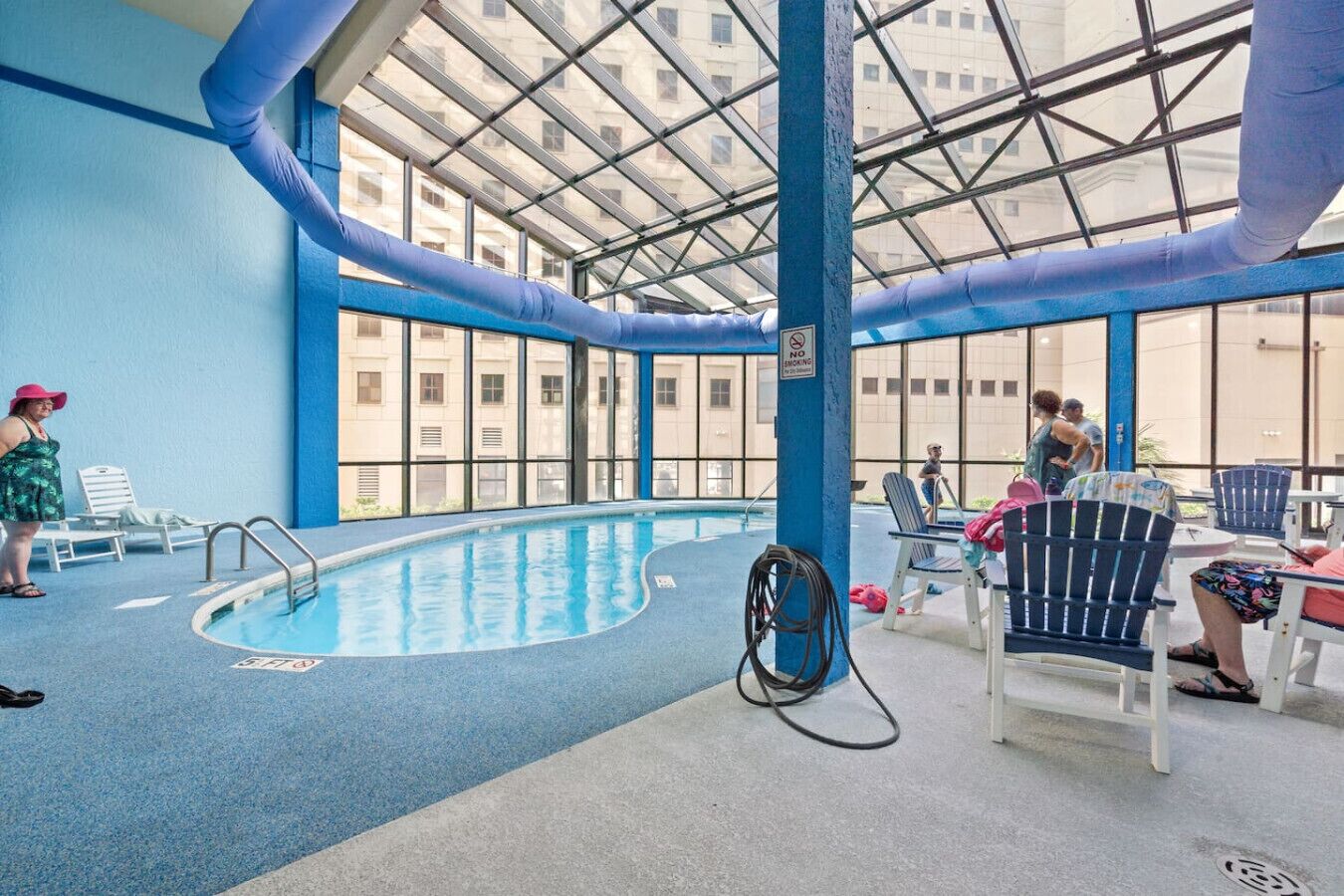 An indoor swimming pool with clear glass ceilings, blue walls, and a poolside area with lounge chairs and tables.