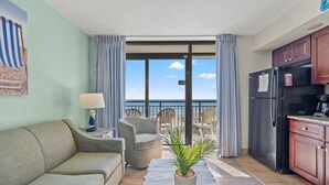 Ett rum med en soffa, stolar, ett litet bord, en krukväxt, en köksdel med skåp och ett kylskåp samt en balkong med utsikt över stranden och havet.