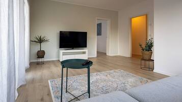 Ein modernes Wohnzimmer mit einem Flachbildfernseher auf einer weißen Konsole, einem grünen Sessel, einem gemusterten Teppich und Holzdielen.