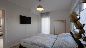 A bedroom with a bed, a TV mounted on the wall, a window with blinds, and a door leading to another room. - Stylish & central vacation apartment in Oranienburg (Oranienburg)