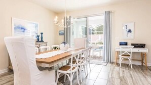 Uma sala de jantar com mesa de madeira e cadeiras brancas, um lustre e vista para o exterior através de uma porta de vidro.