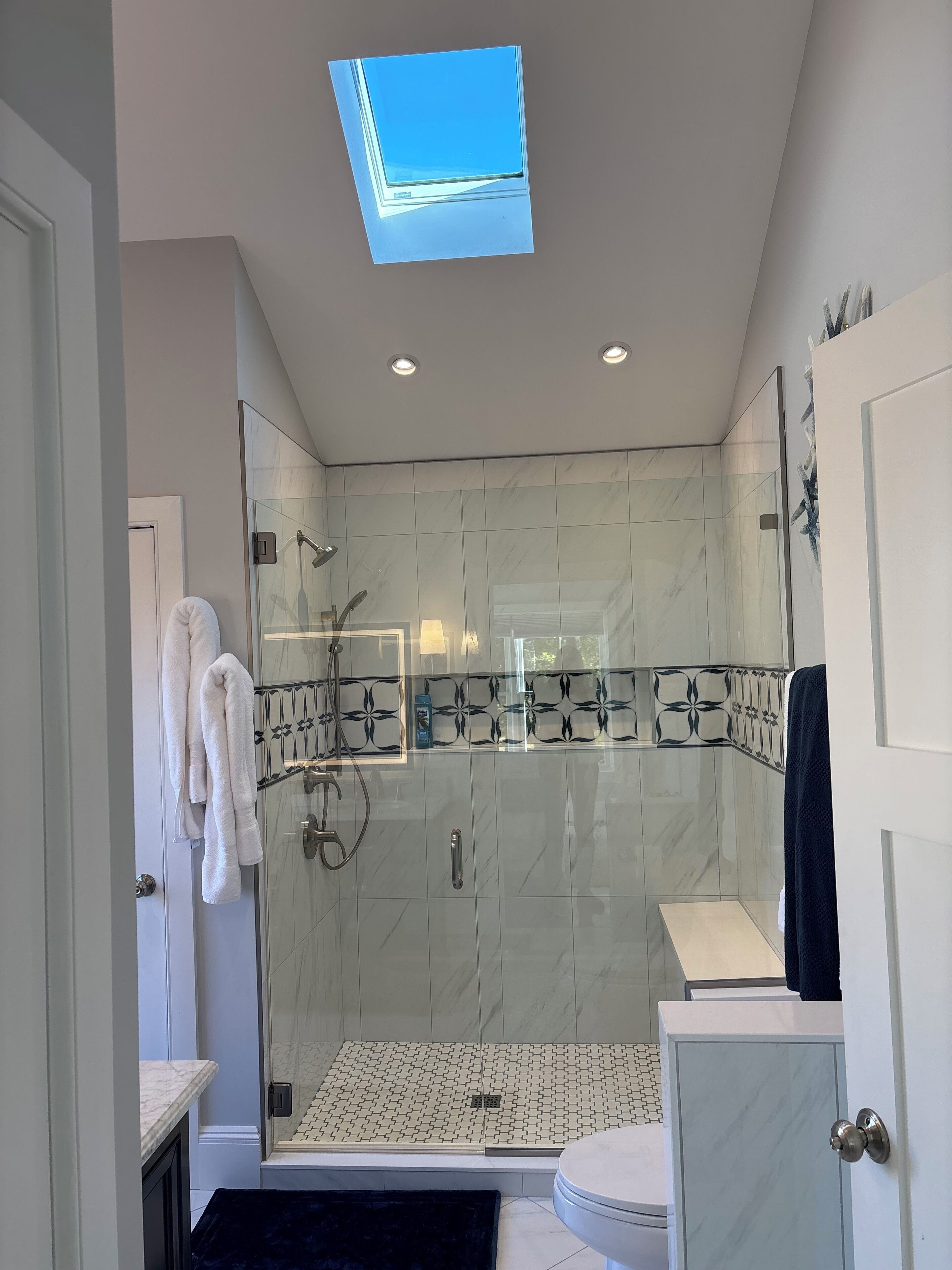 A modern bathroom with a glass-enclosed shower, white marble tiles, and a skylight.