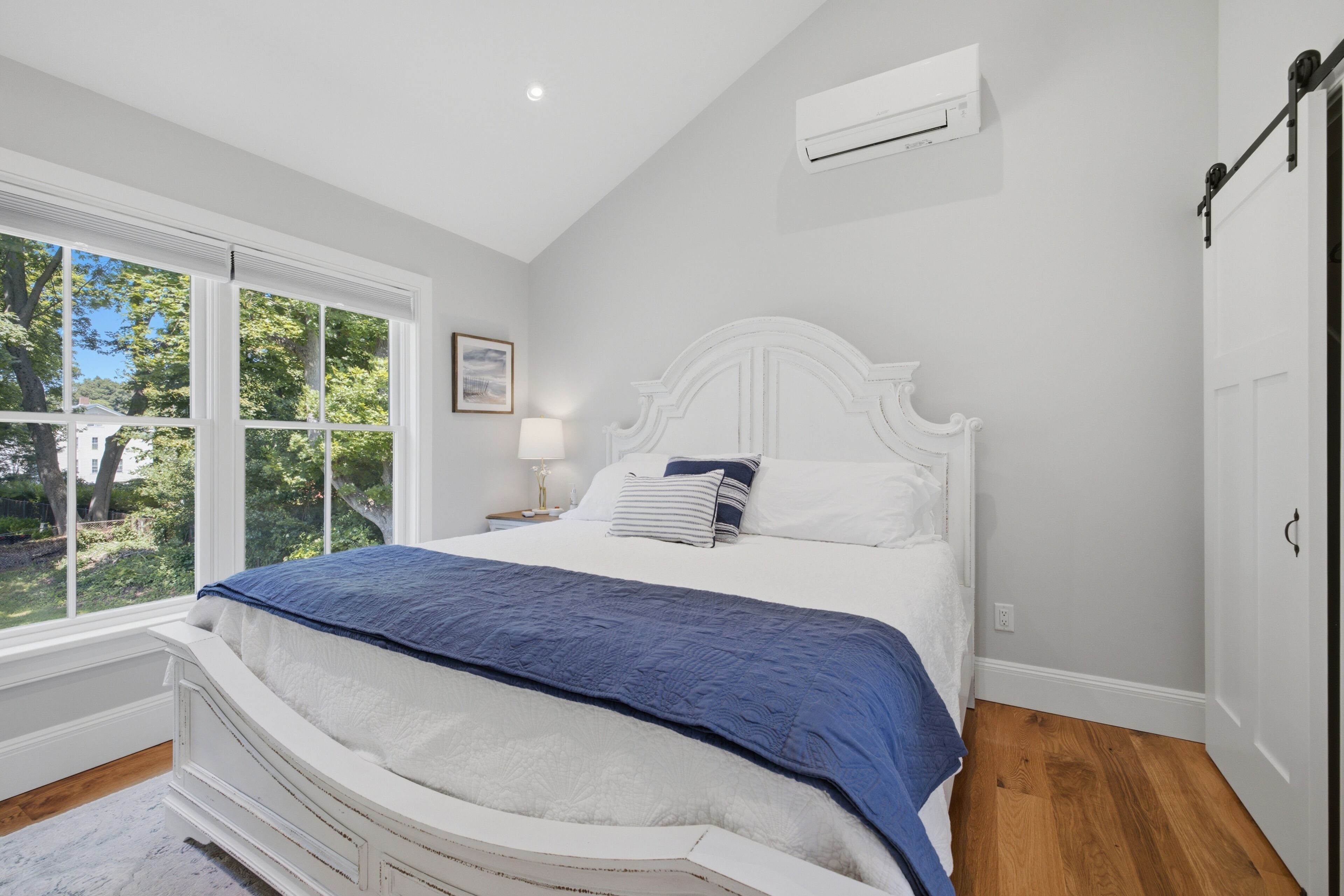 Un dormitorio con un ventanal grande, una cama con cabecera blanca, una mesita de noche y un aire acondicionado instalado en la pared.