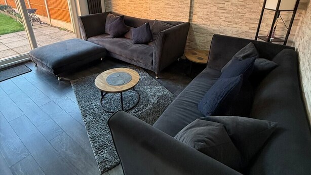 A modern living room with a dark grey sofa, a smaller armchair, and a round coffee table. A large wall clock and a floor lamp are visible. - Luxe Leamington Spa Retreat (Royal Leamington Spa)