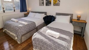A double room with two beds, each with a patterned bedspread and a rolled towel. There are bedside tables with lamps, a window with curtains, and wall art above the beds. - Luxe Leamington Spa Retreat (Royal Leamington Spa)