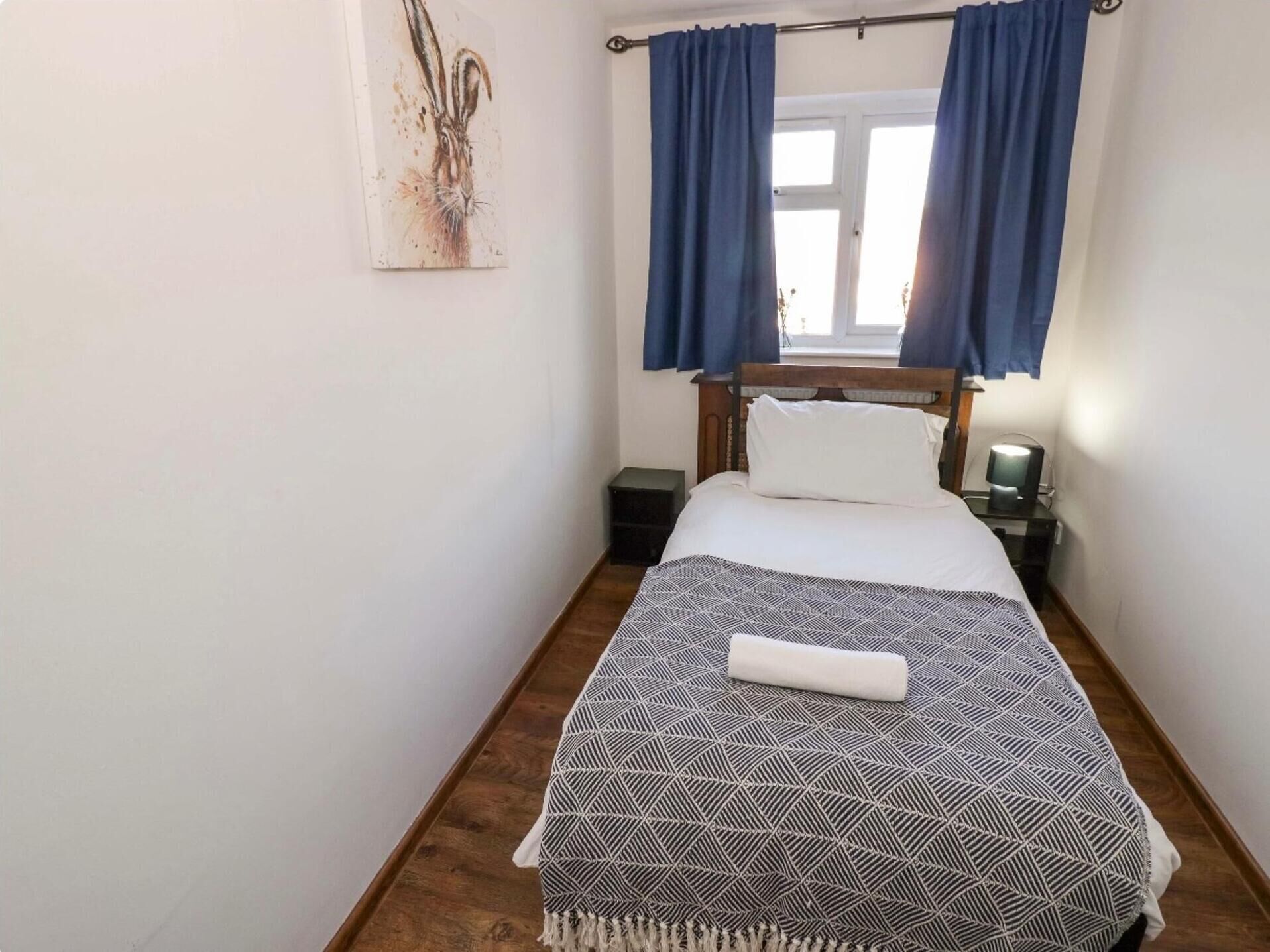 A single bed with a patterned bedspread, a wooden headboard, and a nightstand with a lamp. Blue curtains cover a window, and a painting is on the wall.