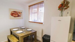 A small kitchen with a wooden table set for four, a white refrigerator, a window with blinds, and a trash bin. - Luxe Leamington Spa Retreat (Royal Leamington Spa)