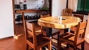 Ein runder Holztisch mit sechs Stühlen, eine Küchenzeile mit weißen Fliesen und Holzregalen sowie ein Fenster mit Vorhang.