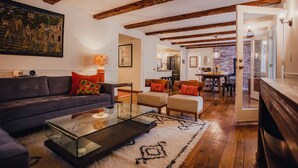 Sala de estar con sofá, sillas, una mesa de centro de vidrio y un tapete. Hay un cuadro en la pared y una cocina al fondo.