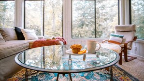 Un cuarto acogedor con una mesa de centro de vidrio, un sofá, una silla y un ventanal con vistas a árboles.