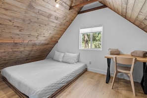Ein Schlafzimmer mit schrägverdeckter Decke, einem Bett, einem Fenster und einem Schreibtisch mit Stuhl.