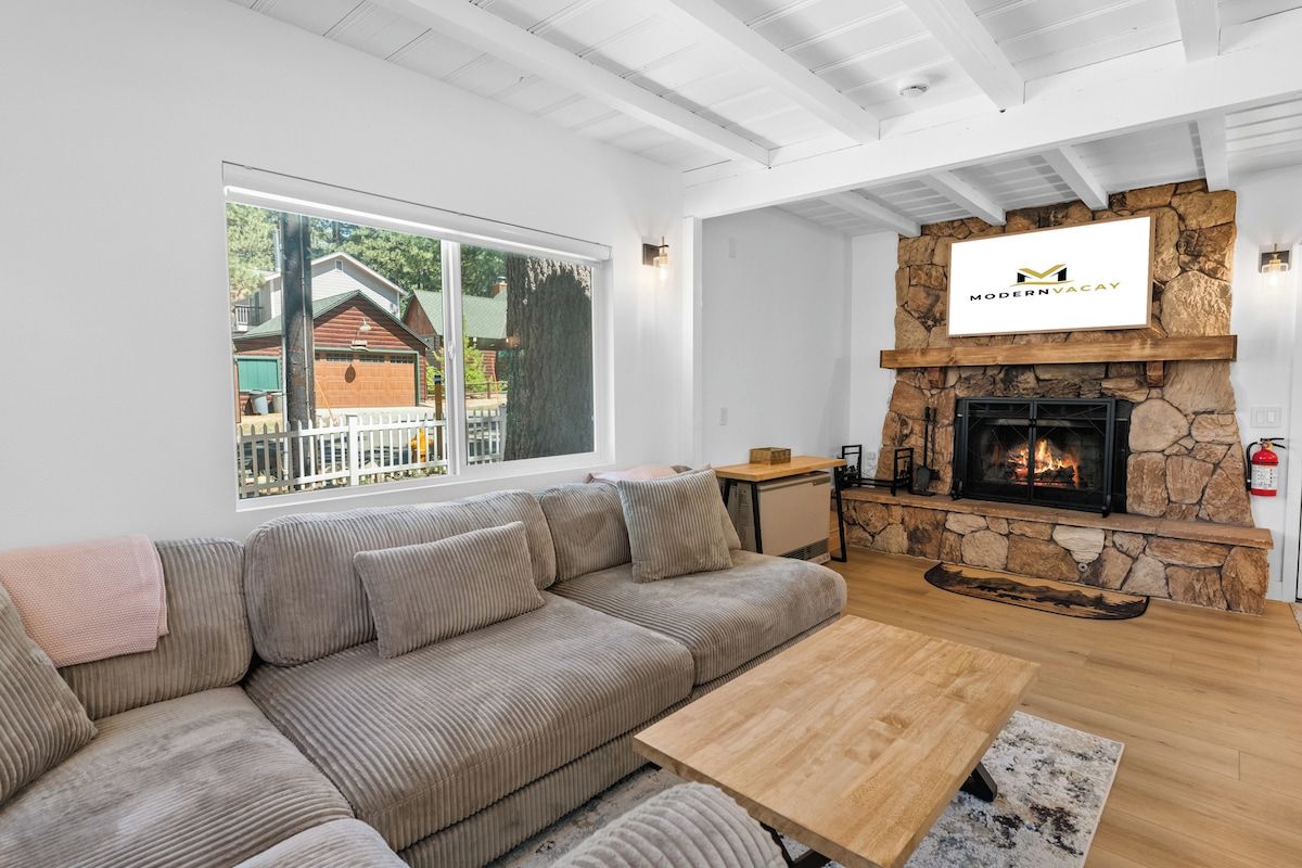 A modern living room with a stone fireplace, a large window, and a view of a house outside.