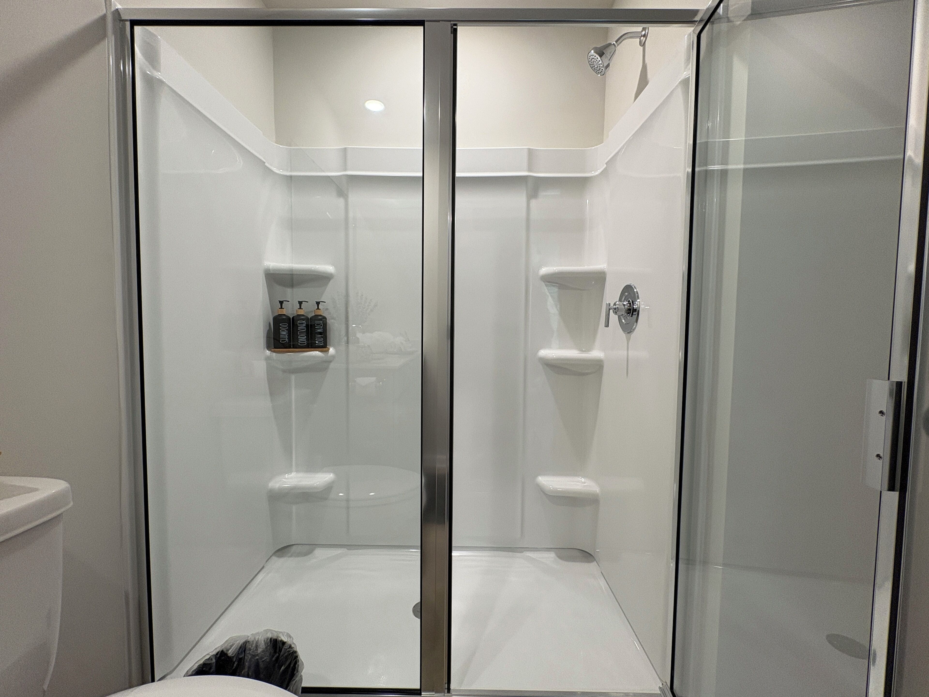 Eine Duschkabine mit Glastüren, ein weiß gefliester Boden, ein Handbrause sowie zwei Flaschen Shampoo und Conditioner auf einem Regal.