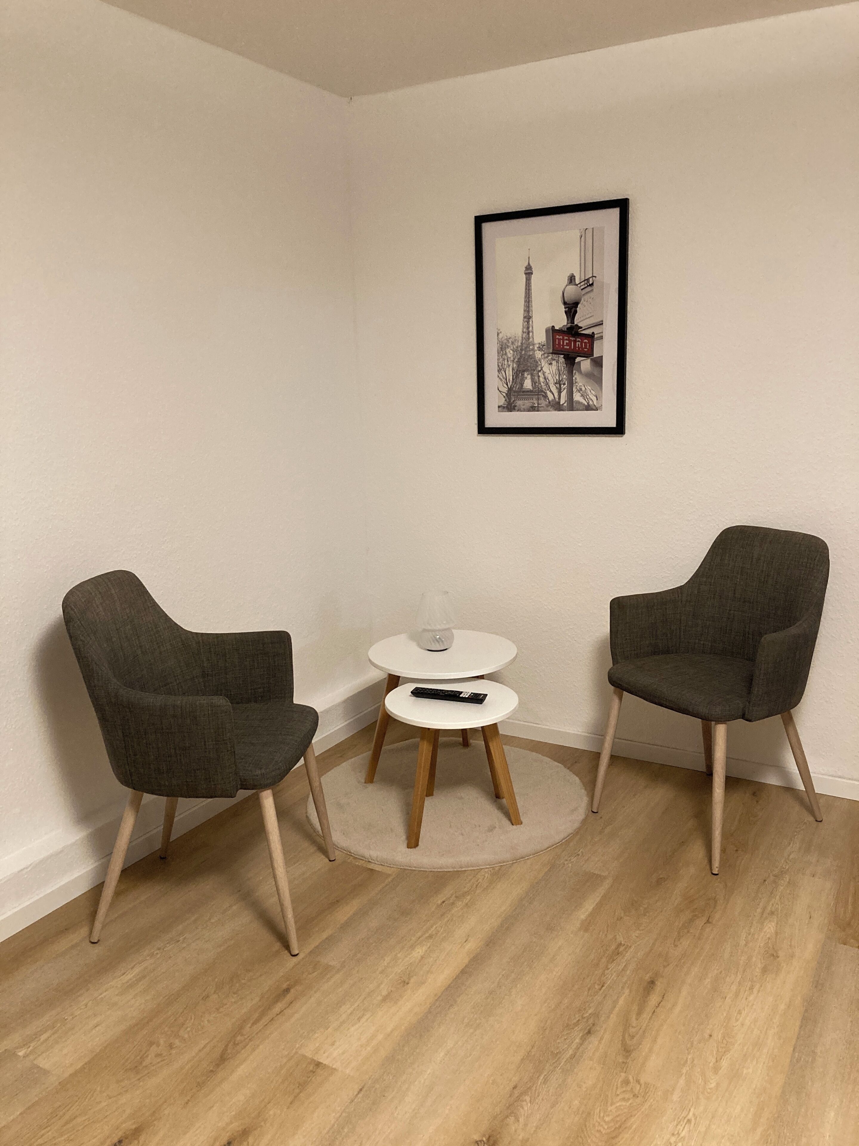 A room with two grey armchairs, a small round table with a white top, and a framed picture of the Eiffel Tower on the wall.