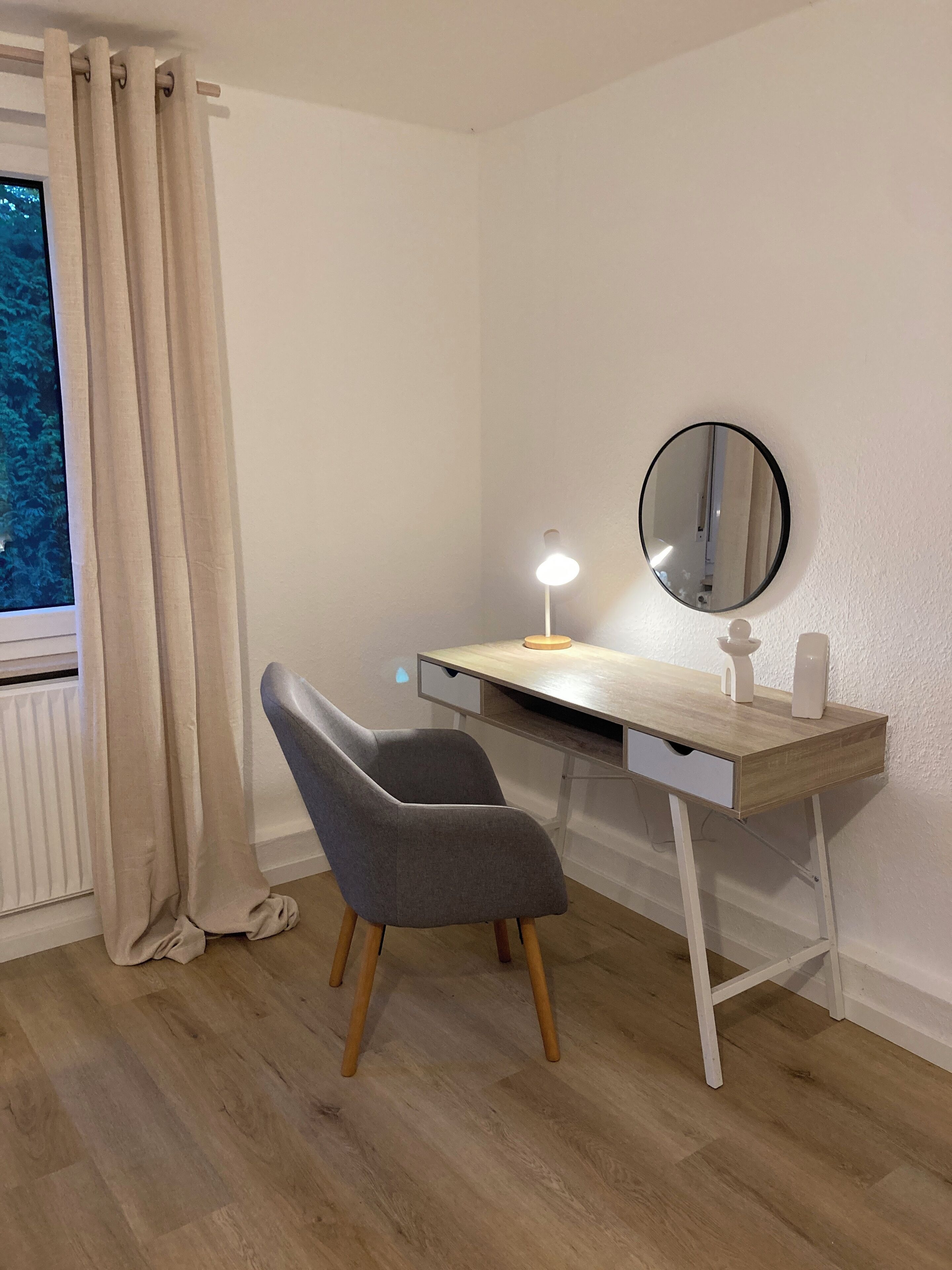 Una sala de estudio moderna con un escritorio de madera, una silla gris, un espejo redondo y una ventana con cortinas.