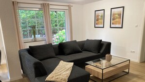 A modern living room with a sectional sofa, a glass coffee table, and a view of greenery through a large window. - Vacation apartment Dorfliebe Minden by Engelmann (Minden)