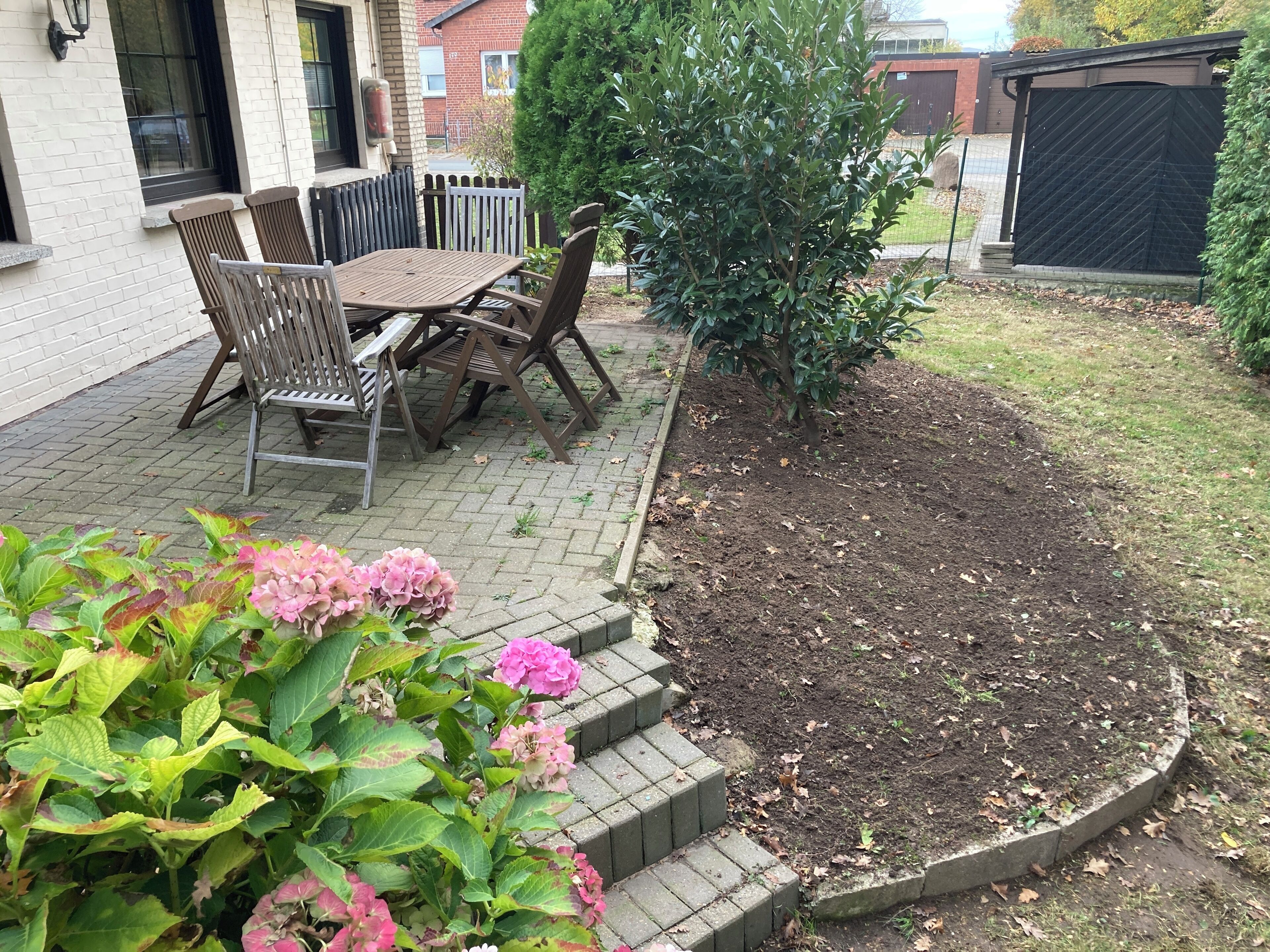 Un patio con una mesa y sillas de madera, rodeado por un borde de ladrillo y un pequeño jardín con flores rosas.