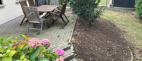 Un patio con una mesa y sillas de madera, rodeado por un borde de ladrillo y un pequeño jardín con flores rosas.