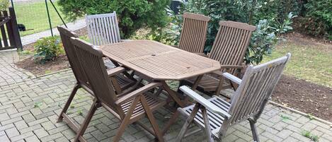 A patio with a wooden table and chairs, surrounded by greenery and a brick pathway.