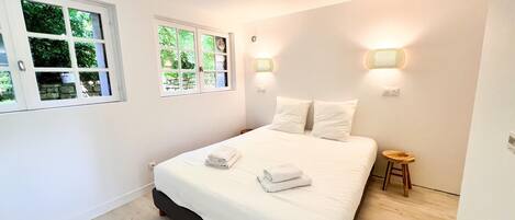 A bedroom with a bed, two bedside lamps, a small wooden stool, and a window with a view of greenery.