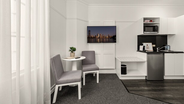 Habitación de hotel compacta y moderna con kitchenette, zona de estar con dos sillas, una mesita y vista panorámica de la ciudad en la pared.