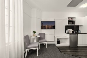 Une chambre d’hôtel moderne et compacte, dotée d’une kitchenette, d’un coin salon avec deux fauteuils, d’une petite table et d’un tableau représentant une vue sur la ville.