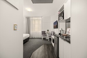Une chambre d’hôtel compacte comprenant un lit, une petite table à manger, une chaise, un coin cuisine et une grande fenêtre avec des rideaux légers.