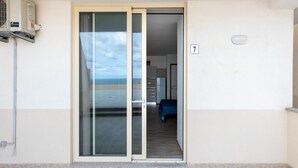 A room with a sliding glass door leading to a balcony, a kitchenette, and a bed with a blue cover. - Aria Condizionata e Terrazza Vista Mare (Castelsardo)