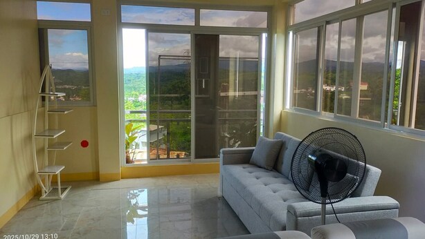 A spacious room with a grey sofa, a standing fan, and a view of greenery through large windows. - Entire rental unit - bright space and a magnificent overlooking view of area. (San Jose del Monte)