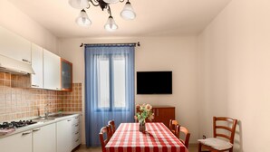 A kitchen with a stove, sink, and cabinets, a dining table with chairs, a blue curtain, and a flat-screen TV on the wall. - 150 Meters From the Sea, 2 Swimming Pools (Valledoria)