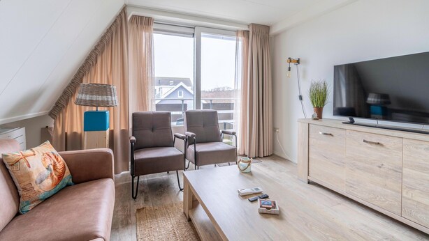 Un salon moderne comprenant un canapé, deux fauteuils, une table basse, un téléviseur posé sur un meuble en bois et une grande fenêtre avec des rideaux.