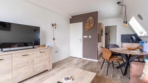 Un salon moderne avec un téléviseur à écran plat, des meubles en bois, une table à manger avec des chaises et, au fond, un coin cuisine.