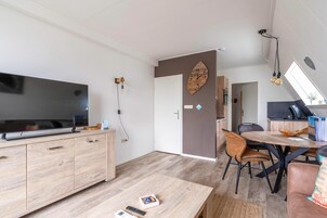 Ein modernes Wohnzimmer mit Fernseher, Holzschränken, einem Esstisch mit Stühlen und einer Küchenzeile im Hintergrund.