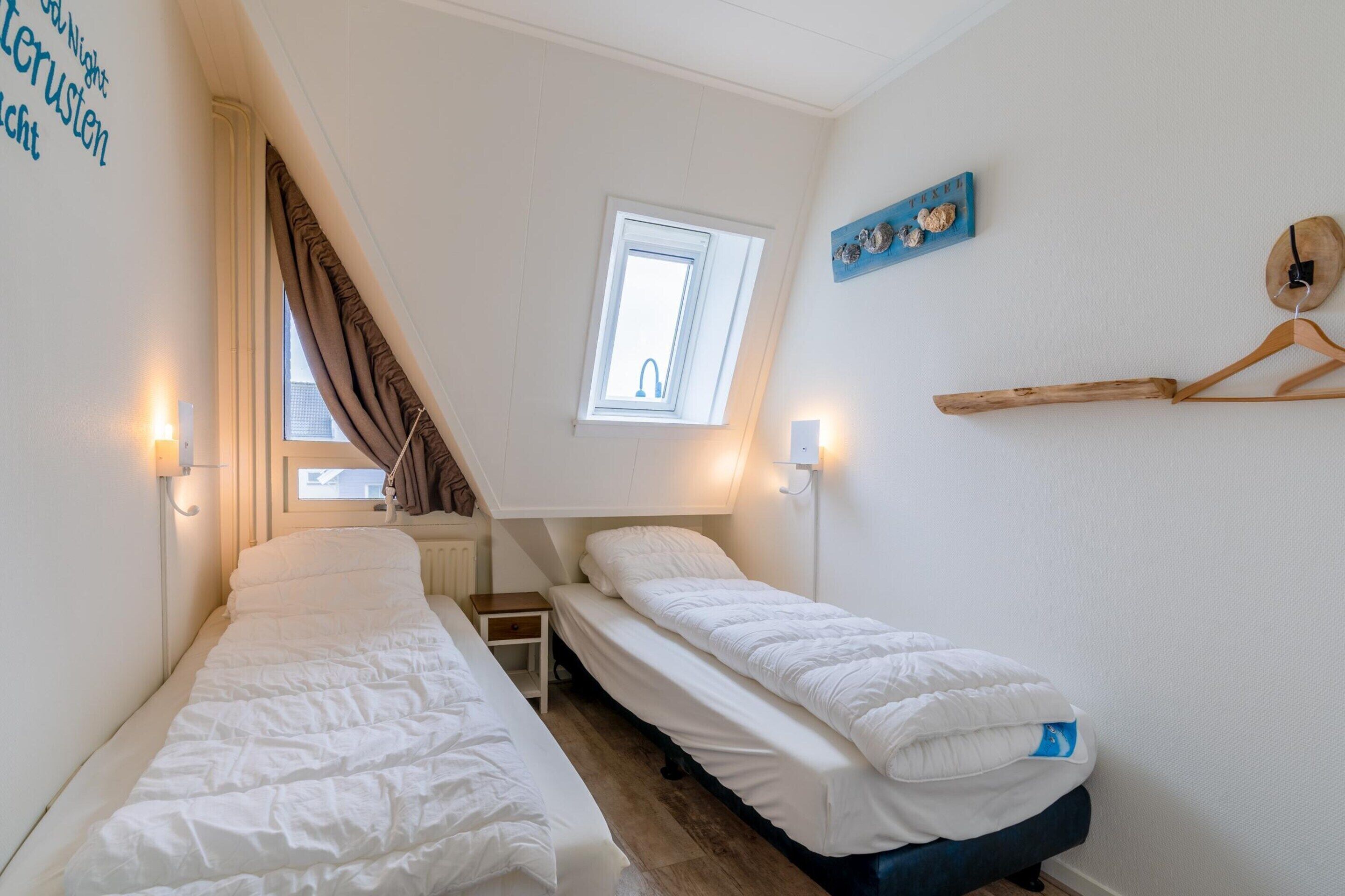 A small, well-lit bedroom with two single beds, a window with curtains, a small table, and a wall-mounted shelf.