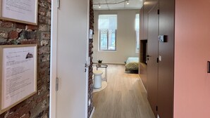 A hallway with a brick wall, a door, a small round table with a plant, a bed, and a window. - Charming Ground-floor Apartment in Bree (Bree)