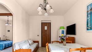 A room with a bed, a sofa, a dining table with chairs, a TV, and a wall-mounted light fixture. - 150 Meters From the Sea, 2 Swimming Pools (Valledoria)