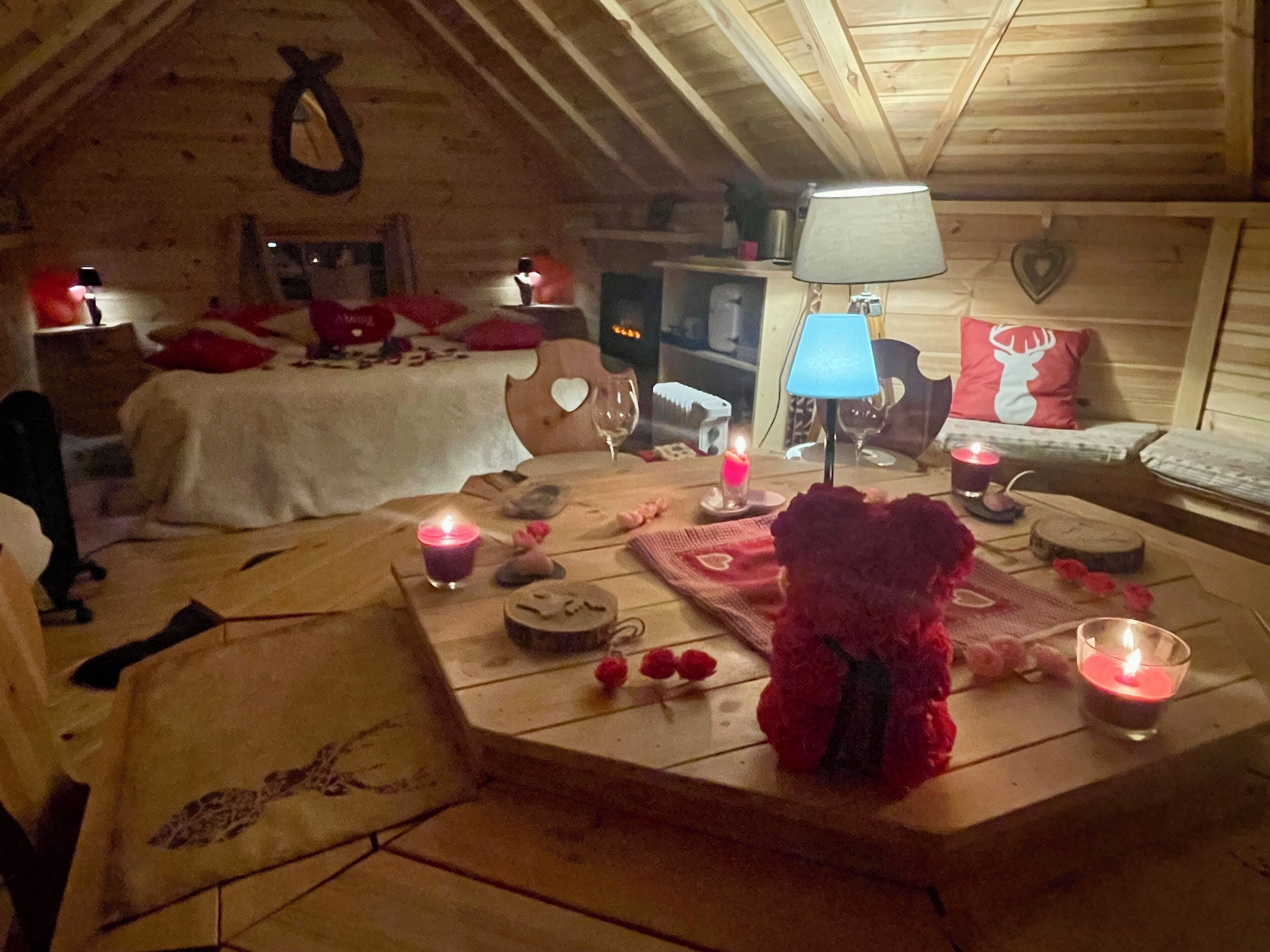 Una habitación acogedora y rústica con techo de madera, una cama con ropa de cama roja, una mesa con velas y un oso de peluche, y una lámpara.