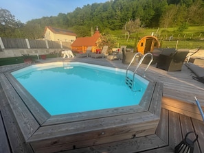 Piscina com deck de madeira, uma espreguiçadeira e um pequeno edifício ao fundo.